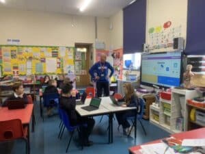 A classroom with children sitting by tables and a man standing in the middle of the room, looking at a screen