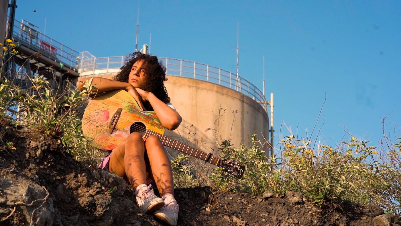 Frances Abigail Bolley sits on a seaside cliff holding a guitar
