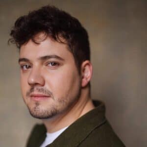 Photo of a person with dark hair and a beard standing in front of  grey wall