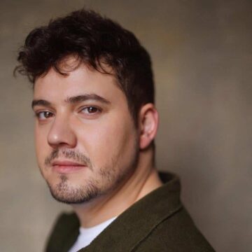 Photo of a person with dark hair and a beard standing in front of  grey wall